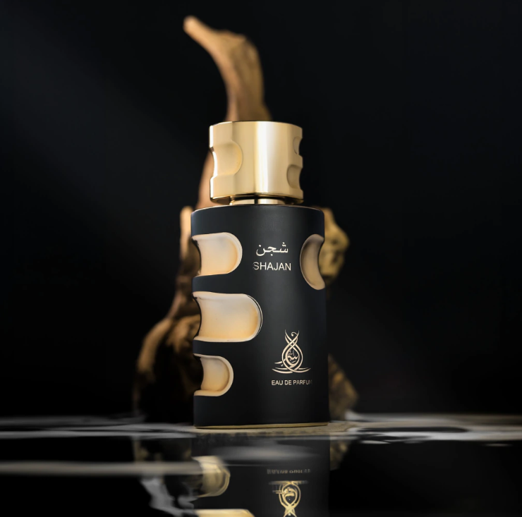 A black cylindrical perfume bottle with a metallic gold cap and gold lettering reading 'SHAJAN' and 'EAU DE PARFUM', shown on a glossy black reflective surface with a blurred gold-toned decorative background. Al Asrar Shajan Feeling Collection Unisex Eau de Parfum 100ml matte black bottle with gold cutout accents dramatic dark editorial shot with golden smoke and reflective surface - Authentic Perfume Store UPC 6292024022906
