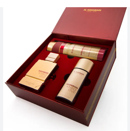 Al Haramain Ambour Oud fragrance gift set in a dark red box, containing a gold perfume bottle, a gold body spray can, and a gold shower gel bottle.