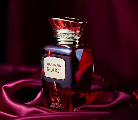 A bottle of Haramain Rouge perfume from the French Collection, featuring a dark red glass body with a metallic cap, resting on draped magenta satin fabric.