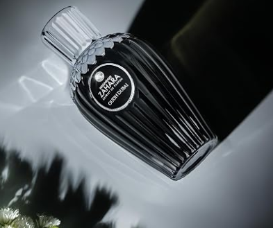 A faceted black glass perfume bottle with a ribbed body and a clear cap, featuring a circular black label with white text that reads 'ZAHRA OUDH DUBAI'. The bottle is shown floating against a dark, moody background with soft light reflections.