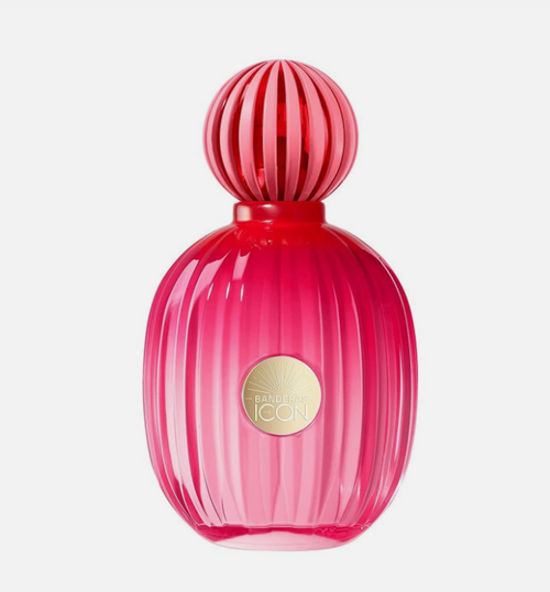 A vibrant pink ribbed glass perfume bottle with a matching spherical cap, featuring a gold circular 'Banderas Icon' label on the front, set against a clean white background.
