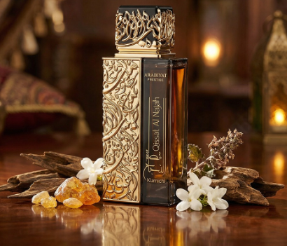 A luxury perfume bottle with ornate gold embossed patterns and a black cap, displayed on a wooden surface with white flowers and amber stones against a warm, softly blurred background.