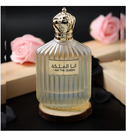 A ribbed clear glass perfume bottle with a gold crown-shaped cap and a beige label reading 'I AM THE QUEEN' in English and Arabic, shown on a dark surface with pink roses and wooden boxes in the background.
