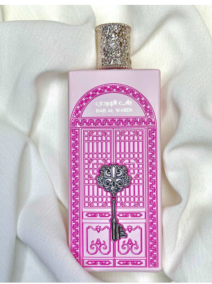 A rectangular perfume bottle with a pink and magenta door illustration design, featuring a silver ornate key motif on the front and a silver patterned cap, set against draped white fabric.