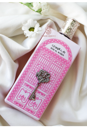 A pink rectangular perfume bottle with a silver ornate key graphic on the front, resting on draped white fabric with a small white flower nearby. The bottle label reads 'Crystal Ball Bath Alchemy' and 'Bath Alchemy' appears as the visible brand.