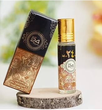 A small cylindrical perfume or fragrance oil bottle with a shiny gold cap stands beside its ornate rectangular box on a rustic wood slice. The packaging is black and gold with decorative swirls, and the bottle label shows the number 24.