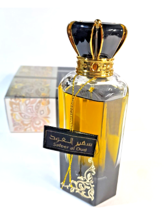 A clear glass perfume bottle with a black and gold crown-shaped cap, filled with amber-colored fragrance. A black hanging tag reads 'شهر العود / Safier al Oud' in Arabic and English.