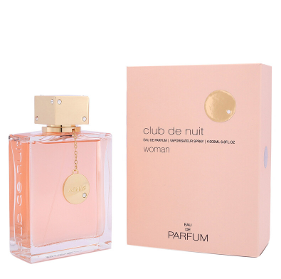A bottle of Club de Nuit Woman Eau de Parfum next to its light pink packaging box, both featuring gold accents and a soft pink color, on a white background.