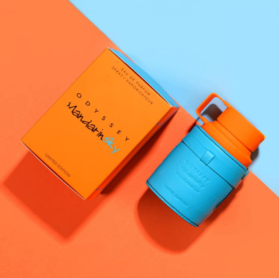 A blue and orange Odyssey Wander-in-Any plastic water bottle with a matching orange lid and carrying loop, shown next to its orange product box on a diagonally split orange and light blue background.