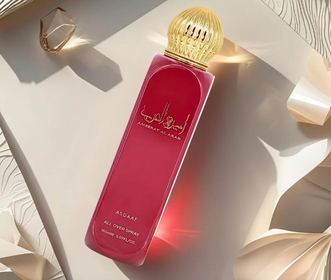 A rectangular pink perfume bottle with a ribbed gold cap, photographed on a cream-colored surface with soft shadows and decorative paper elements. The bottle has gold Arabic script and the brand name 'ASDAAF' visible on the front.