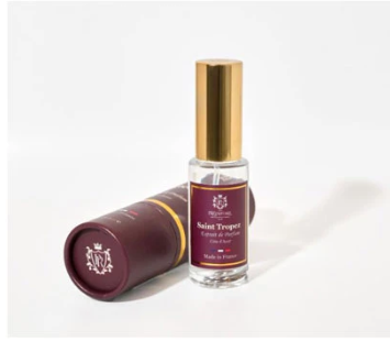 A small clear glass perfume bottle with a gold cap stands upright next to a dark burgundy cylindrical tube on a clean white background. The bottle label reads Saint Tretor.