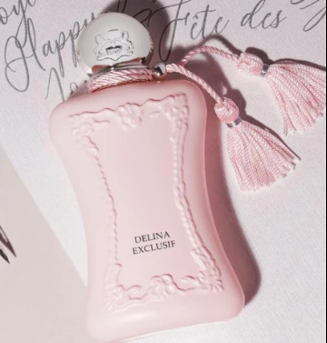 A pale pink perfume bottle with embossed floral detailing and a white spherical cap, shown on a light surface with decorative script text and pink tassels.