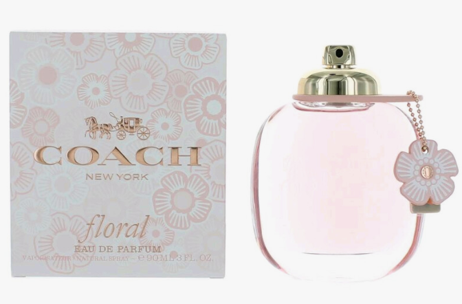 Coach New York Floral Eau de Parfum bottle next to its patterned packaging box. The clear glass bottle contains a light pink liquid and features a gold spray cap, a small flower-shaped charm, and a pink floral pattern on the box.