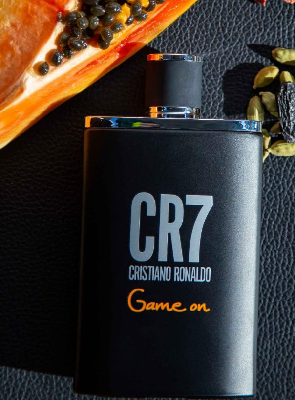 A black rectangular bottle of CR7 Cristiano Ronaldo Game on fragrance with silver and orange lettering, photographed on a dark textured surface with sliced papaya and cardamom seeds nearby.