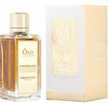 A bottle of Édou Américaine Assemblage Signature perfume with amber liquid and a gold cap, shown next to its white and gold packaging box.