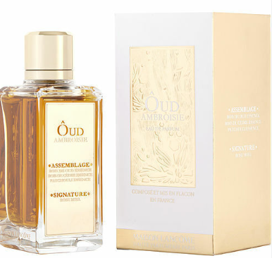 A bottle of Édou Américaine Assemblage Signature perfume with amber liquid and a gold cap, shown next to its white and gold packaging box.