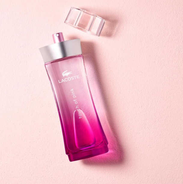 Lacoste Touch of Pink perfume bottle in a vibrant pink gradient glass with a silver cap, shown with its matching cap off on a textured light pink background.