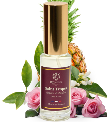 A clear glass perfume bottle with a metallic gold cap and a dark burgundy label reading Saint Tropez, surrounded by pink roses, green leaves, and a pineapple on a white background.