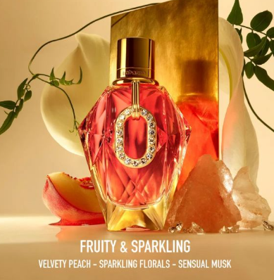A faceted pink-red perfume bottle with a gold cap and a jeweled O-shaped detail on the front, styled with peach slices, white flowers, green leaves, and crystals. Text at the bottom reads 'FRUITY & SPARKLING VELVETY PEACH - SPARKLING FLORALS - SENSUAL MUSK'.