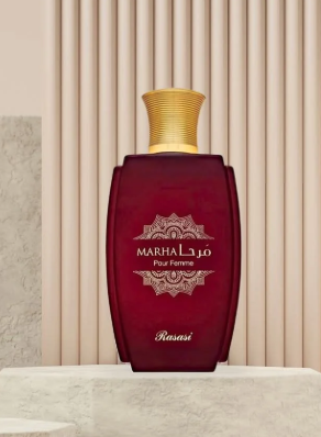 A bottle of MARHA Pour Femme perfume, featuring a deep burgundy red body with a textured gold cap, displayed on a marble surface against a beige paneled background.