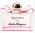 A bottle of Salvatore Ferragamo Signorina in fiore perfume, featuring a clear glass bottle filled with light pink liquid, a metallic rose gold cap, and a pink ribbon bow, set against a plain white background.