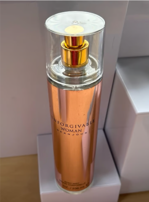 A bottle of Dean John 'Unforgivable Woman' body spray with a metallic rose gold body, gold pump, and clear cap, standing on a white display block.