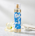 A bottle of Tommy Bahama Set Sail St. Barts body mist stands on a white marble surface with a white hibiscus flower and folded white towels beside it. The bottle has a blue and white tropical floral label with gold accents and a clear cap.