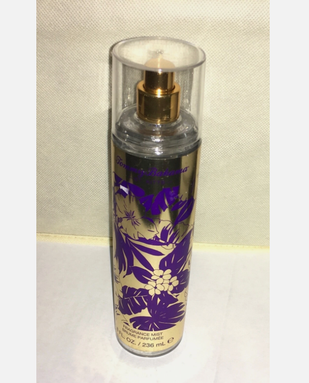 A cylindrical perfume spray bottle with a clear cap and gold pump, featuring a gold label with purple floral graphics and text, set against a light beige fabric background.