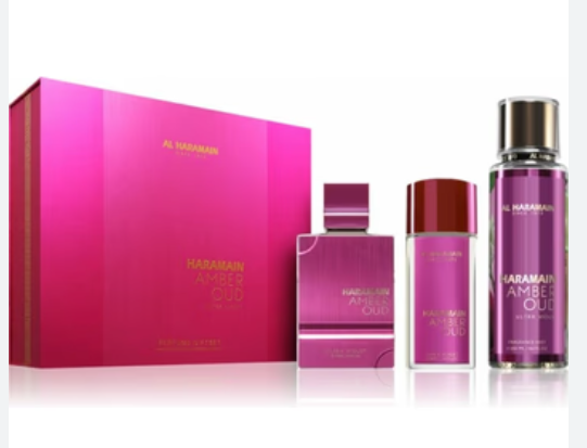 Al Taraiman Haramain Amber Oud perfume set featuring a large pink gift box, a rectangular spray bottle, a cylindrical bottle with a red cap, and a tall cylindrical bottle with a silver cap, all in shades of pink and purple on a white background.