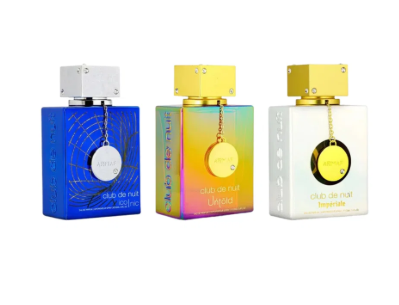 Three perfume bottles from the brand Clâm de Nuit, labeled Club de Nuit Impériale, shown side by side against a white background. The bottles feature iridescent, metallic, and solid color designs in blue, yellow, and white with gold caps.