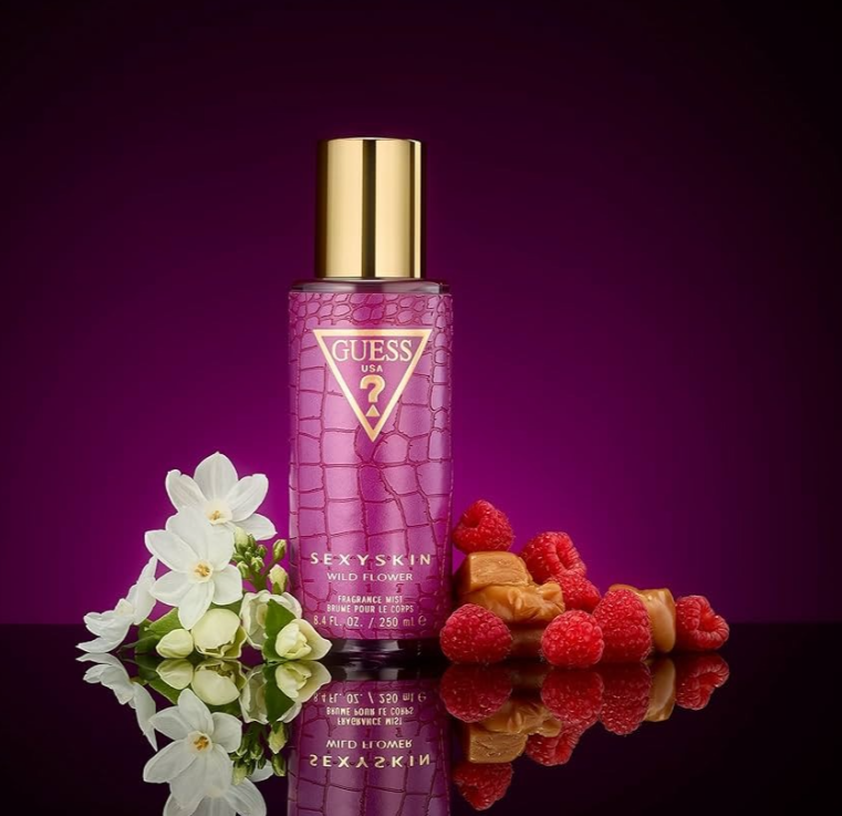 Guess Sexyskin Fragrance Oil bottle in a purple crocodile-embossed texture with a gold cap, styled with white flowers and raspberries on a reflective black surface against a dark purple gradient background.