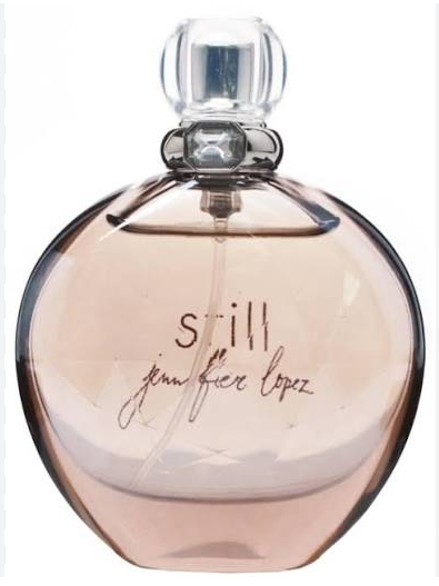 A round glass perfume bottle with a clear cap and a pale pink liquid inside, featuring the text 'still jennifer lopez' printed on the front.