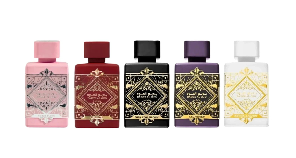 Five ornate perfume bottles lined up against a white background, each with a different body color and matching cap: pink, deep red, black, purple, and white, all decorated with intricate gold geometric labels.
