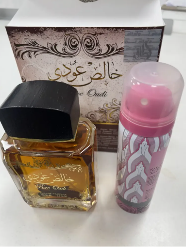 A bottle of Pure Oudi perfume with amber liquid and a black cap, next to a pink spray canister, with the branded box behind them on a white surface.