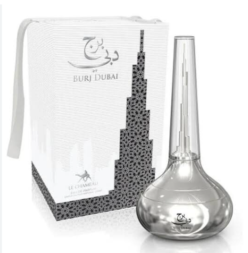 A clear glass perfume bottle with a tall neck and rounded base, shown beside its white and black patterned box. The box features Arabic script and the text 'Buri Dubai' and 'Le Chemise'.