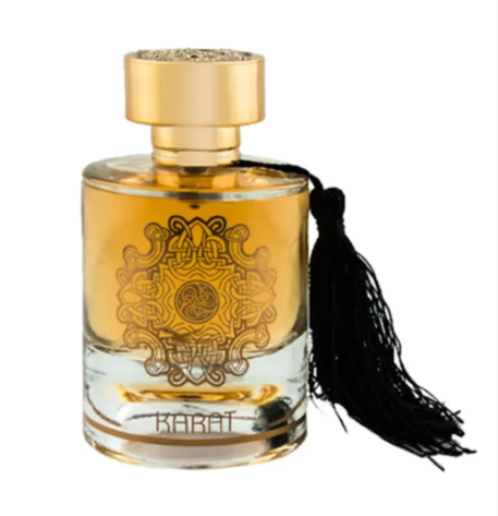 A clear glass perfume bottle filled with amber-gold liquid, featuring an ornate gold emblem on the front, a gold cap, and a long black tassel hanging from the side. The word 'KABAT' is printed near the bottom.