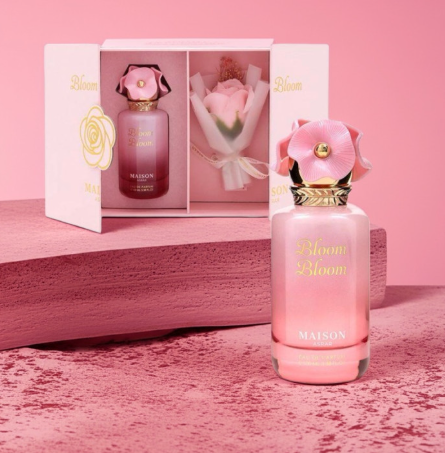 Maison Bloom Eau de Parfum bottle in pink with a flower-shaped cap, shown in front of its matching pink gift box and a small wrapped rose on a pink background.