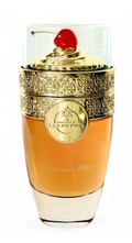 A luxury perfume bottle with a clear glass body, filled with amber-colored liquid, featuring an ornate gold metal band and a lid topped with a red cherry, on a white background.