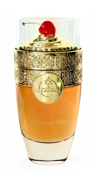 A luxury perfume bottle with a clear glass body, filled with amber-colored liquid, featuring an ornate gold metal band and a lid topped with a red cherry, on a white background.